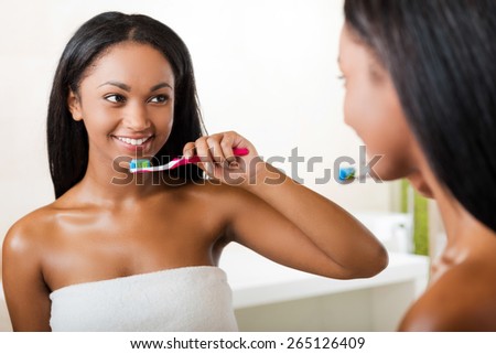 Taking care of her teeth. Beautiful young African woman brushing her teeth and smiling while standing against a mirror in bathroom