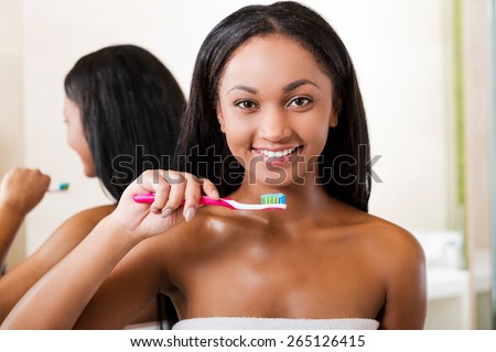 Teeth care. Beautiful young African woman brushing her teeth and smiling while standing against a mirror in bathroom