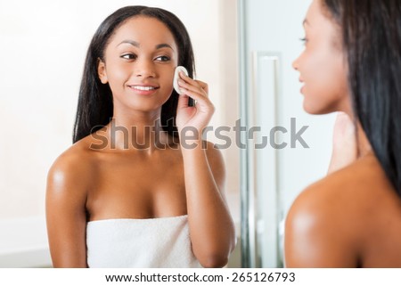 Taking good care of her face. Beautiful young African woman cleaning her face with sponge and smiling while standing against a mirror in bathroom