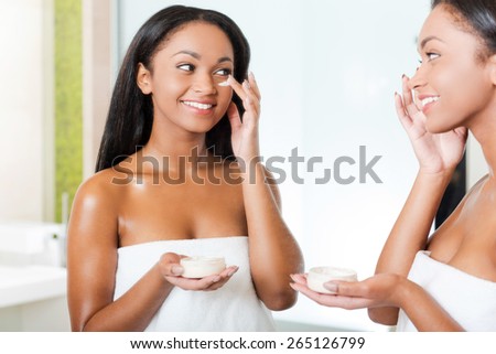 Taking good care of her skin. Beautiful young African woman spreading cream on her face and smiling while standing against a mirror in bathroom