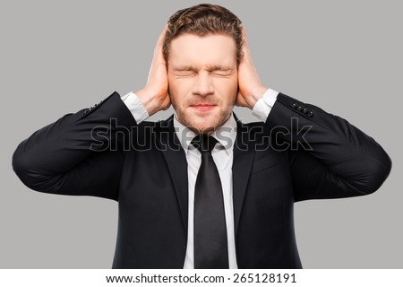 This is too loud! Frustrated young man in formalwear holding head in hands and keeping eyes closed while standing against grey background