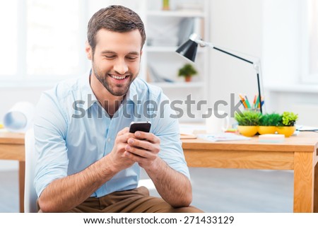 Staying in touch with his colleagues. Handsome young man holding mobile phone and smiling while sitting at his working place