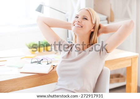 Satisfied with the work done. Beautiful young woman holding hands behind head and smiling while sitting at her working place in office