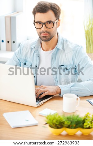 Working on creative project. Handsome young man in shirt and eyewear working on laptop while sitting at his working place