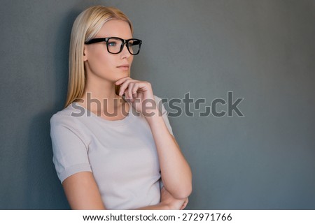 Lost in thoughts. Thoughtful young blond hair woman holding hand on chin and looking away while standing against grey background