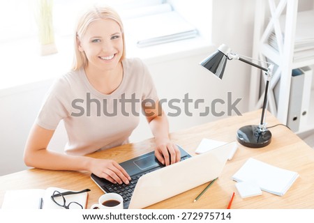 Enjoying her work. Top view of beautiful young woman looking at camera and smiling while sitting at her working place in office