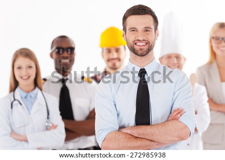 When I grow up I will be a businessman. Confident young man in shirt and tie keeping arms crossed and smiling while group of people in different professions standing in the background