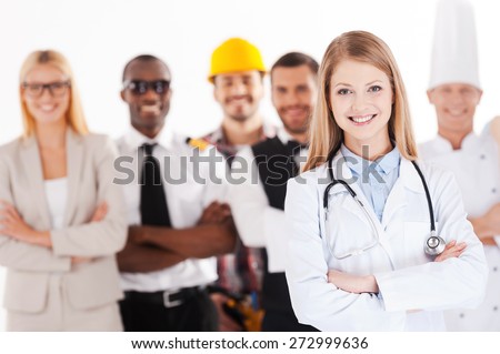 When I grow up I will be a doctor. Beautiful young female doctor keeping arms crossed and smiling while group of people in different professions standing in the background
