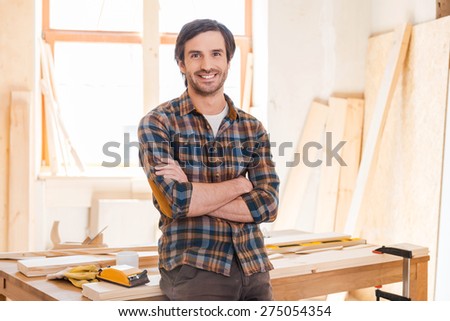 My workshop is my world! Happy young male carpenter keeping arms crossed and leaning at the wooden table with diverse working tools laying on it