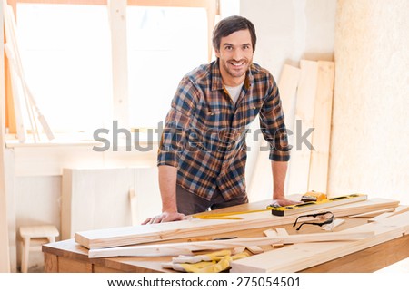 Smiling woodworker. Cheerful young male carpenter leaning at the wooden table with diverse working tools laying on it