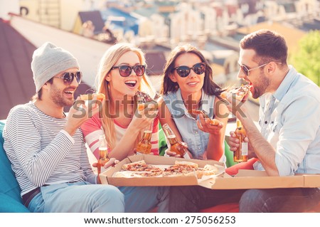 Friends and pizza. Four young cheerful people eating pizza and drinking beer while sitting at the bean bags on the roof of the building