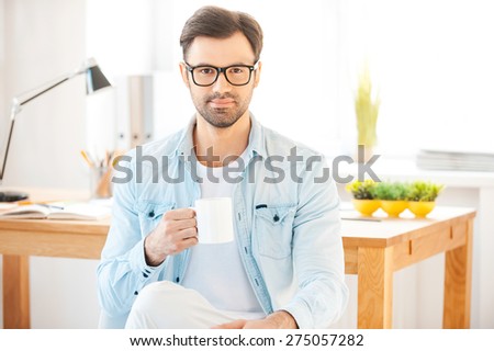 Cup of coffee for great ideas. Handsome young man in shirt and eyewear holding cup of coffee and looking at camera while sitting in front of his working place
