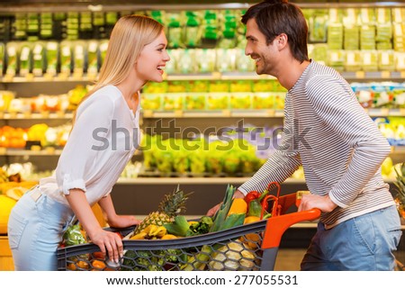 Finding joy in the simplest things. Cheerful young couple leaning at the shopping cart and looking at each other while shopping in a food store