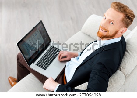 Working with pleasure. Top view of happy bearded businessman working on laptop and smiling at camera while sitting on sofa
