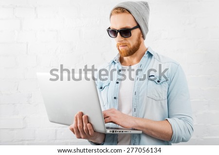Smart technology for perfect style. Concentrated young bearded man holding laptop and working on it while standing against brick wall