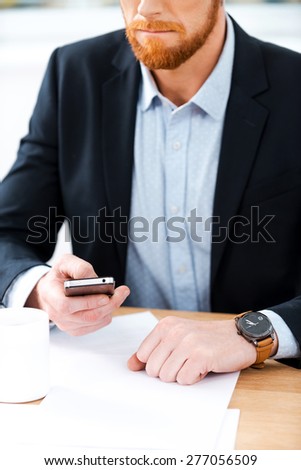 Busy working. Close-up of bearded businessman wearing watch and holding mobile phone while sitting at his working place