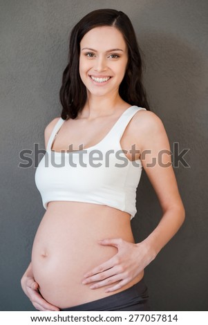 Enjoying her special period. Beautiful pregnant woman holding hand on her abdomen and touching her face while standing against white background