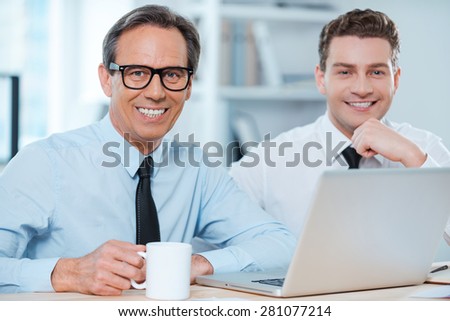 Two professionals at work.Two cheerful business people in formalwear working on laptop and smiling at camera while sitting at working place