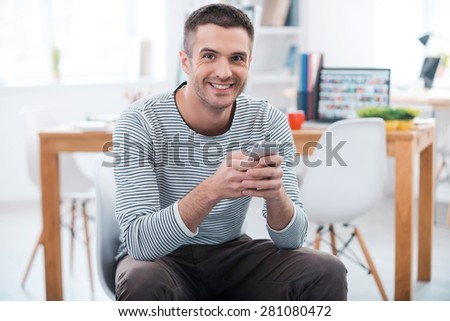 Young and successful. Happy young man holding mobile phone and looking at camera while sitting in the office