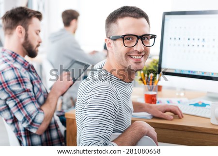 We keep it casual in our office. Happy young man looking over shoulder and smiling while sitting at desk with his colleagues working in the background