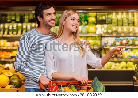 Look at that! Beautiful young woman pointing away and smiling while standing close to her boyfriend in food store