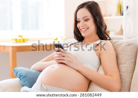 Surrounded by positive emotions. Happy pregnant young woman holding mobile phone and smiling while sitting on sofa