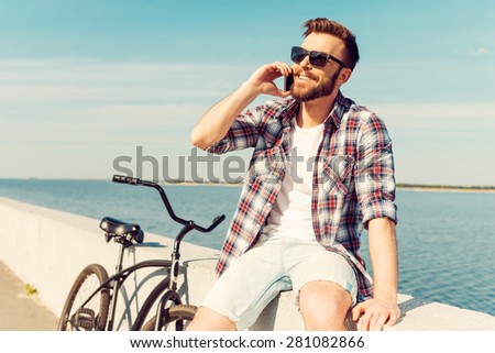 Perfect day for cycling! Cheerful young man talking on the mobile phone and smiling while sitting on parapet near his bicycle