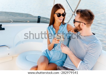 Celebrating their special anniversary. Smiling young couple holding glasses with champagne and looking at each other while sitting on the board of yacht