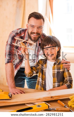 Turning their visions into reality. Cheerful young male carpenter embracing his son while leaning at the wooden table with diverse working tools laying on it