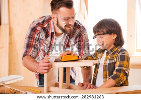 Carpentry team. Cheerful young male carpenter helping his son tosand wooden chair in workshop
