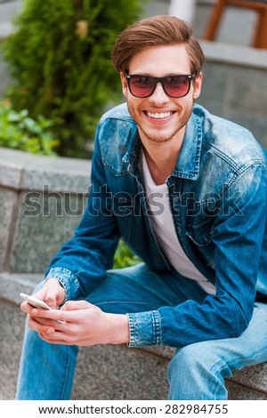 Cheerful man outdoors. Smiling young man holding mobile phone and looking at camera while sitting outdoors