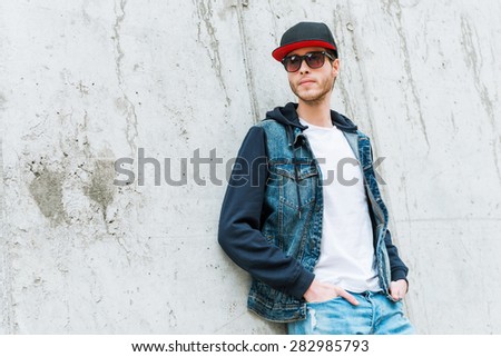 Cool man in cap. Confident young man in headwear holding hands in pockets while leaning at the concrete wall