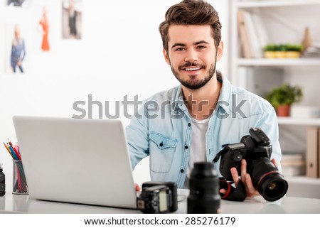 My job is my passion. Handsome young man holding camera and smiling while sitting at his working place