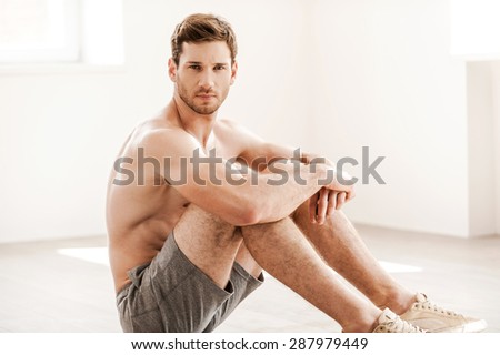 Rugged and handsome. Handsome young shirtless man in shorts sitting on the floor and looking at camera