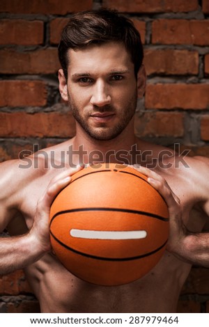He is willing to this game. Handsome young muscular man holding basketball ball while standing against brick wall