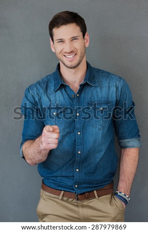 You can do it! Happy young man pointing at camera and smiling while standing against grey background