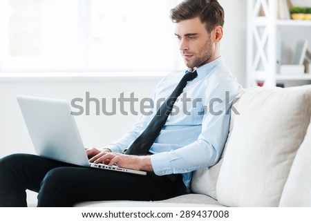Working with comfort. Serious young businessman working on laptop while sitting on sofa