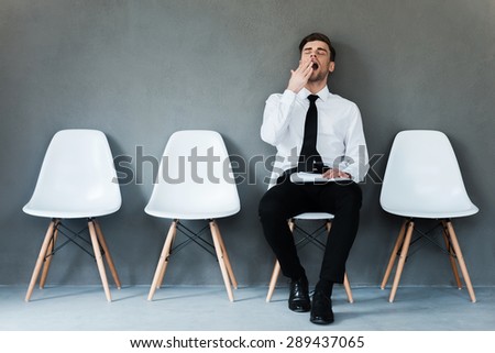 Tired of waiting. Tired young businessman holding paper and yawning while sitting on chair against grey background