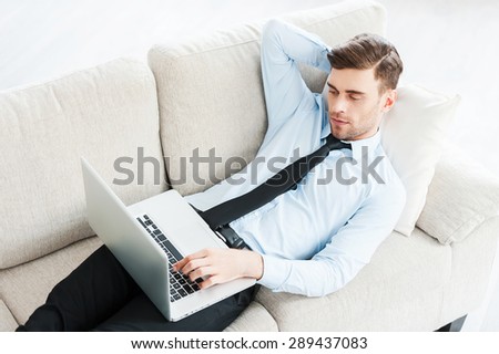 Working at home. Top view of young businessman working on laptop and holding hand behind head while lying on sofa