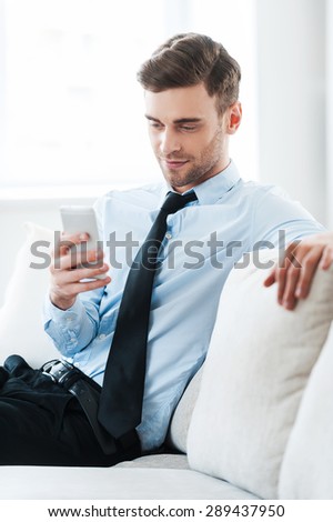 Feeling comfortable while doing business. Smiling young businessman holding mobile phone while sitting on sofa