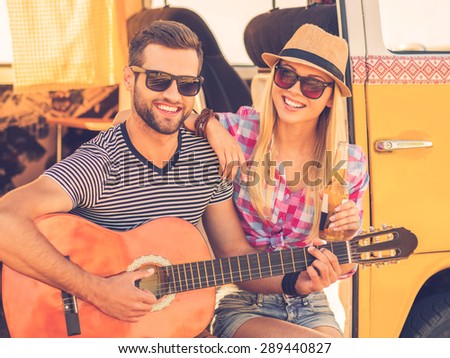 Music break during road trip. Handsome young man sitting in minivan and playing guitar while his cheerful girlfriend bonding to him and smiling