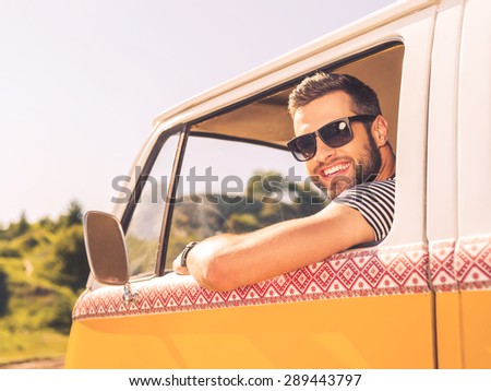 In searching of adventures. Happy young man looking at camera and smiling while sitting at the front seat of his minivan