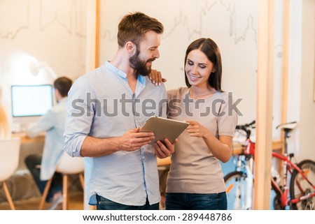 Just look at this! Cheerful young man and woman discussing something and looking at digital tablet together while their colleagues working in the background