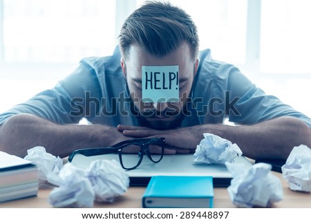 He needs help. Hopeless young beard man with adhesive note on his forehead leaning at the desk and keeping eyes closed while sitting at his working place in office