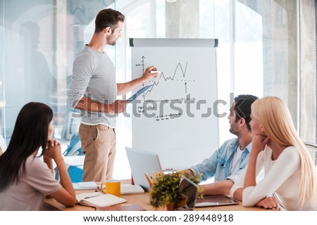 Discussing company progress. Confident young man standing near whiteboard and sketching while his colleagues sitting at the desk