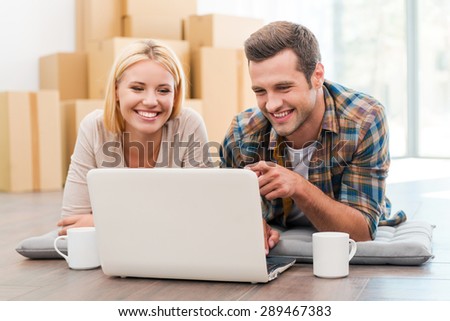 In search of a good moving company. Cheerful young couple laying on the floor of their new apartment and looking at laptop while cardboard boxes laying in the background