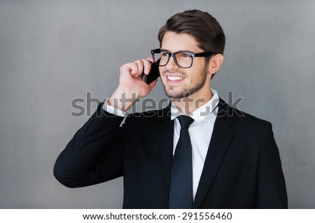 Good business talk. Happy young businessman talking on the mobile phone and looking away while standing against grey background