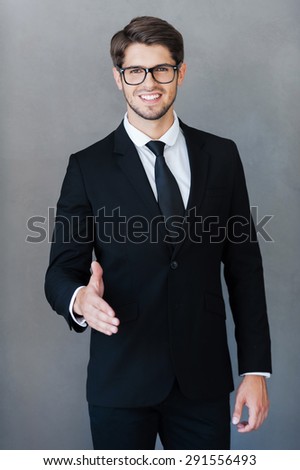 Nice to meet you at our company! Cheerful young businessman stretching out hand for shaking while standing against grey background