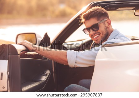 Always ready for good driving. Cheerful young man smiling at camera and closing the door while sitting inside of his white convertible