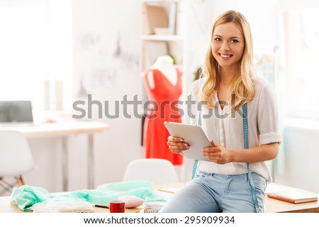 Confident fashion expert. Beautiful young woman holding digital tablet and smiling while sitting on the desk in her fashion workshop